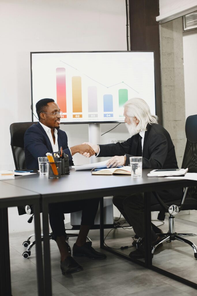 Two diverse businessmen in suits shaking hands over a desk in a modern office.